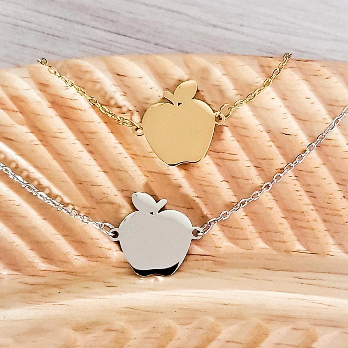 Two apple-shaped pendants on gold and silver chains on a wooden surface
