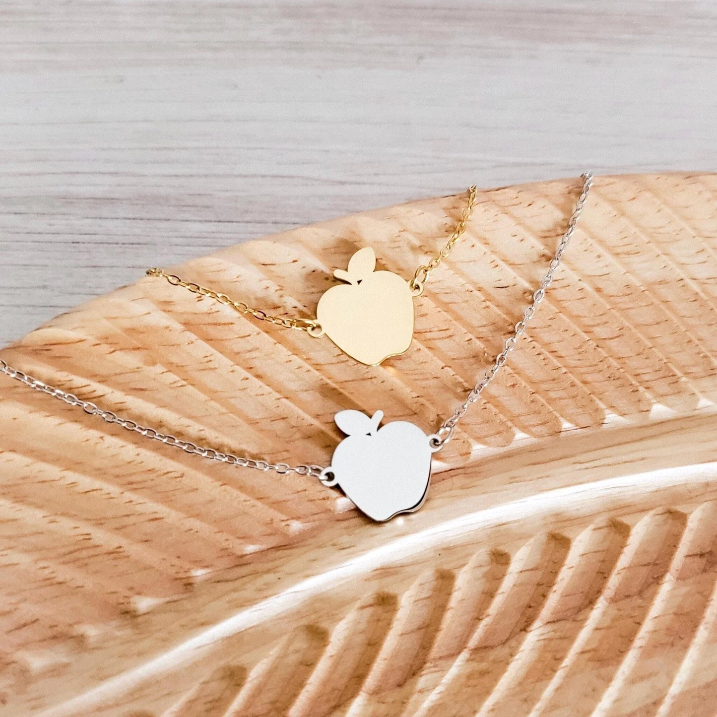 Two apple-shaped pendants on gold and silver chains on a wooden surface