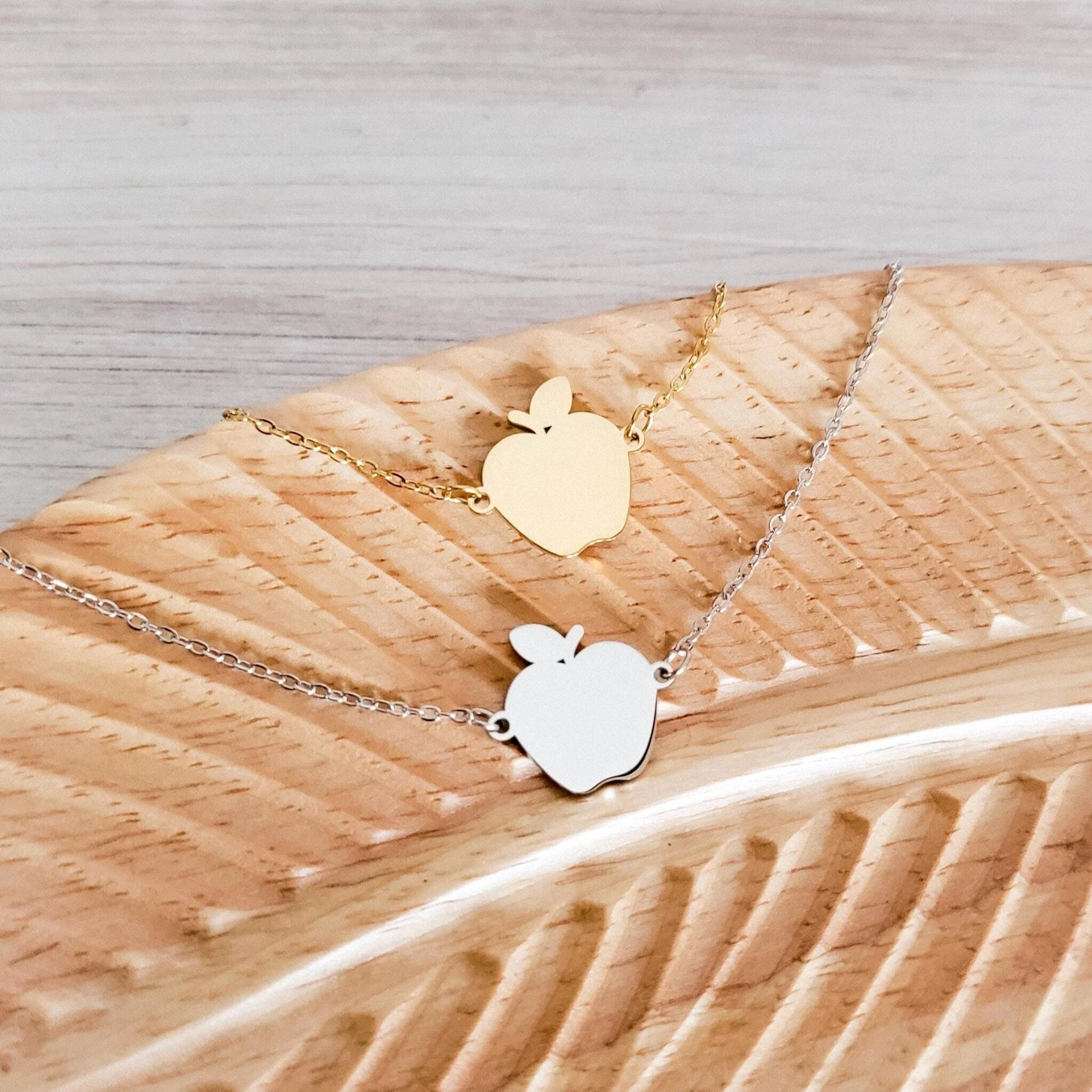Two apple-shaped pendants on gold and silver chains on a wooden surface