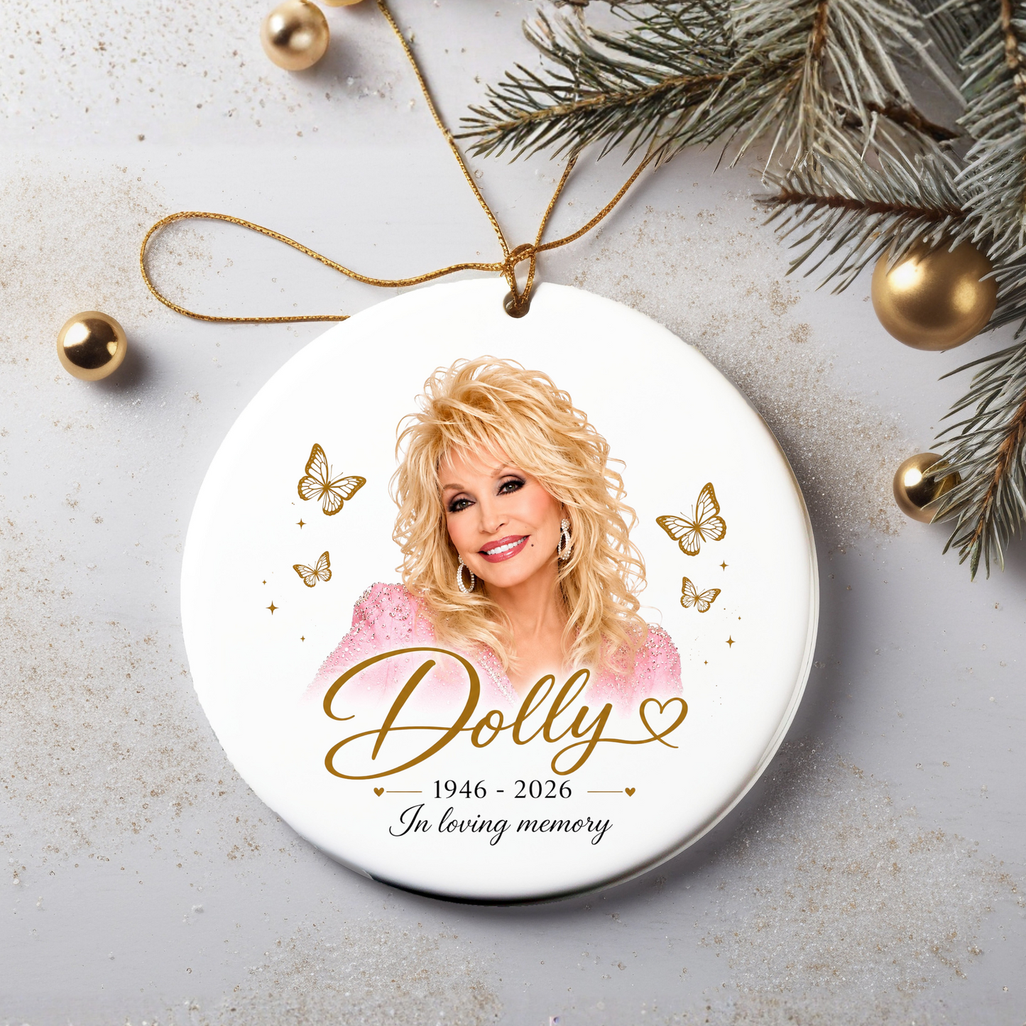 A round white ceramic Christmas ornament featuring a portrait of Dolly Parton with gold script text reading 'Dolly 1946-2026 In loving memory,' surrounded by gold butterflies and stars. The ornament hangs from a gold string and is styled on a light gray surface with pine branches, gold ornaments, and glitter.