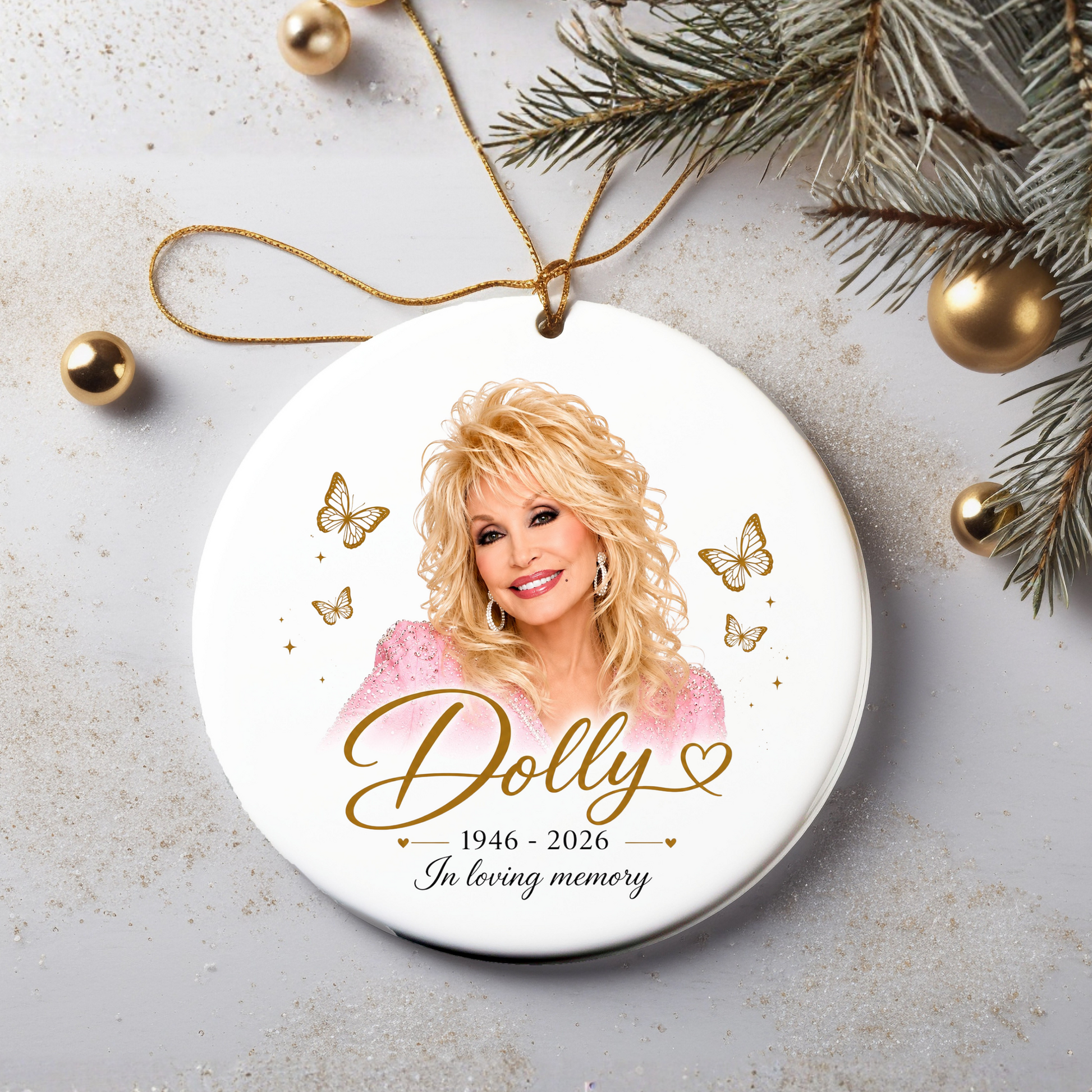 A round white ceramic Christmas ornament featuring a portrait of Dolly Parton with gold script text reading 'Dolly 1946-2026 In loving memory,' surrounded by gold butterflies and stars. The ornament hangs from a gold string and is styled on a light gray surface with pine branches, gold ornaments, and glitter.