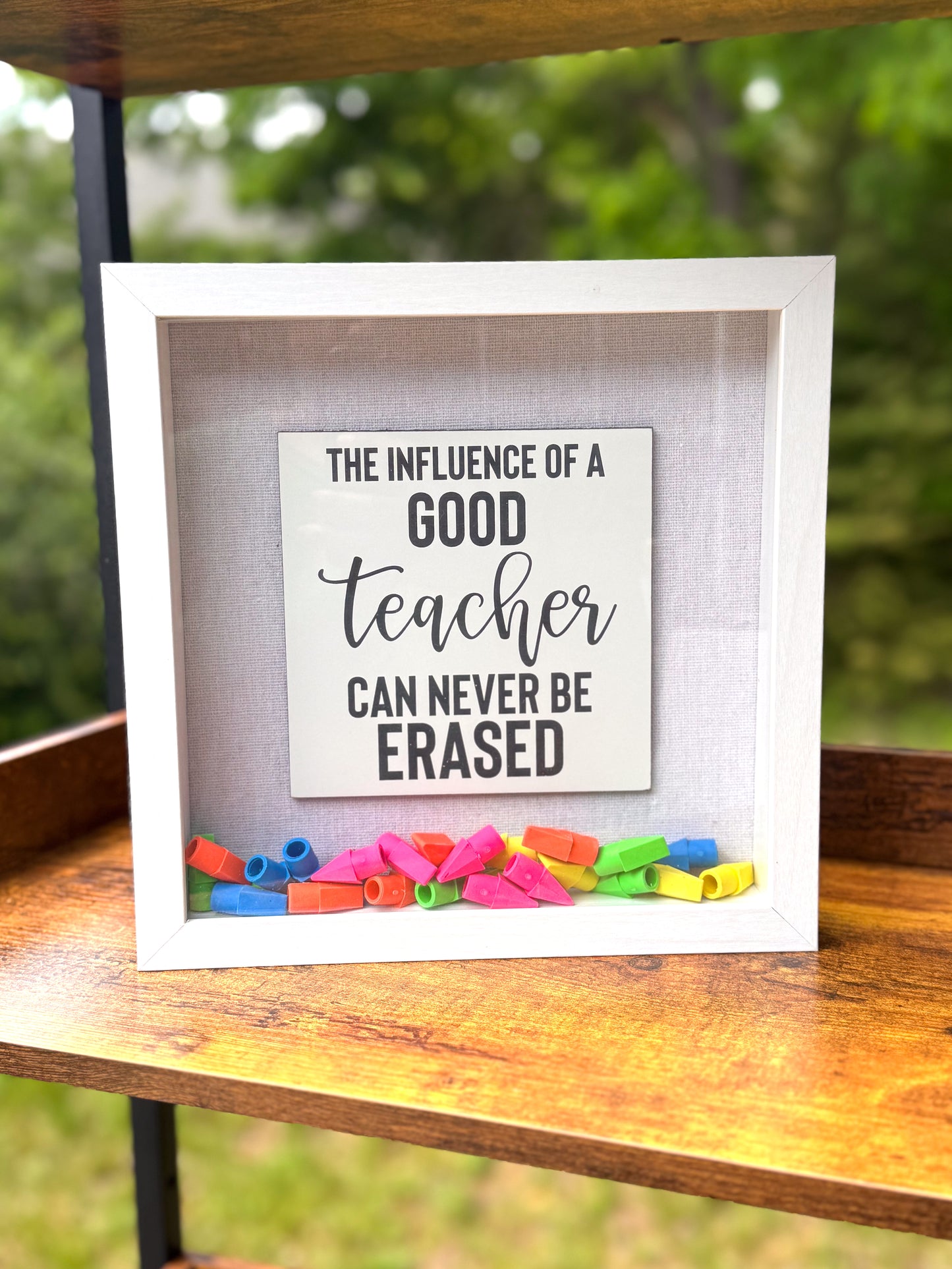 Framed sign with 'The influence of a good teacher can never be erased' quote on a wooden surface with colorful erasers.