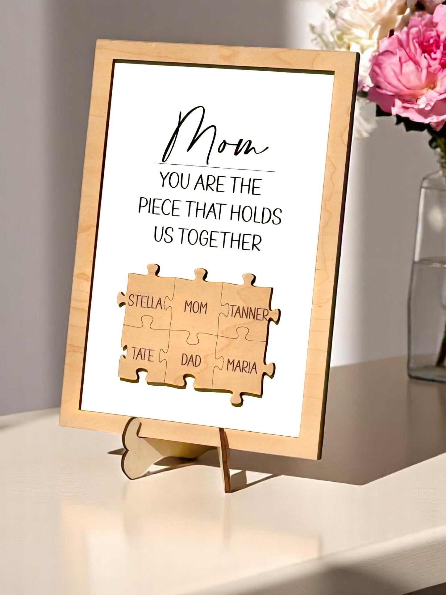 A personalized wooden puzzle frame with family names, displaying a message to 'Mom' that she is the piece that holds us together, placed on a wooden stand. Puzzle pieces with Mom in the center surrounded by husband and children names