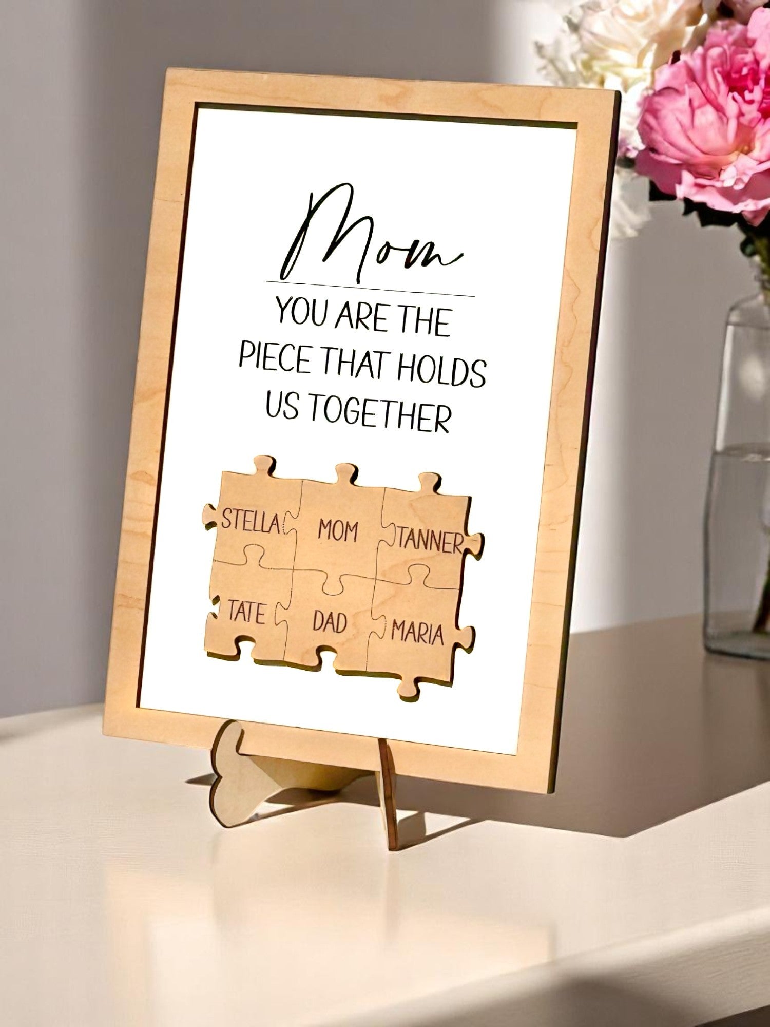 A personalized wooden puzzle frame with family names, displaying a message to 'Mom' that she is the piece that holds us together, placed on a wooden stand. Puzzle pieces with Mom in the center surrounded by husband and children names