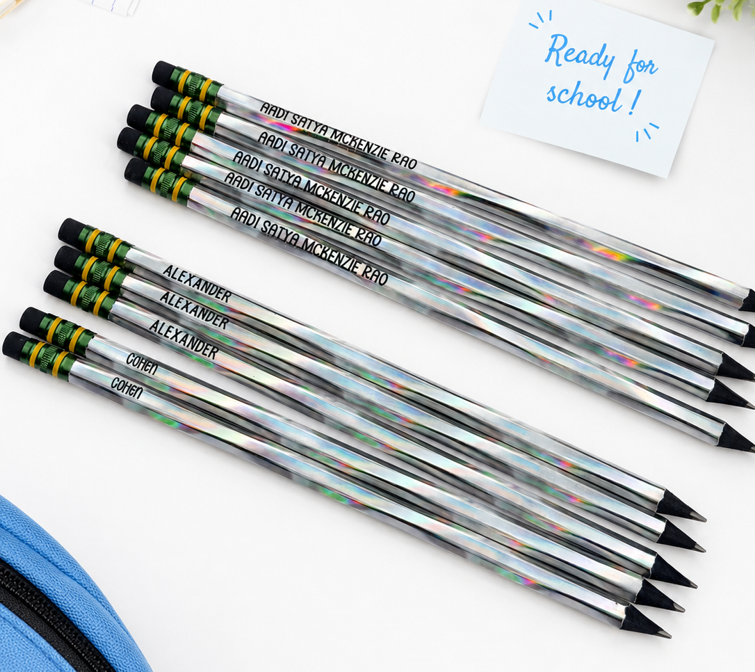 Six personalized school pencils with holographic silver barrels and black erasers, arranged on a white desk alongside a blue pencil case, a notebook, a small plant, and a note that says 'Ready for school!'.