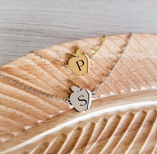 Two gold apple-shaped pendants on a chain necklace for teachers against a wooden surface.
