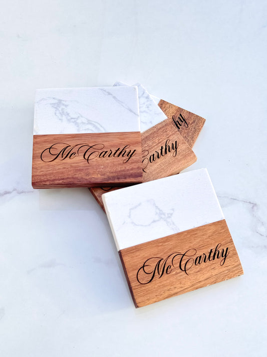 A set of personalized marble and acacia wood coasters with the name 'McCarthy' engraved on the wooden holder.