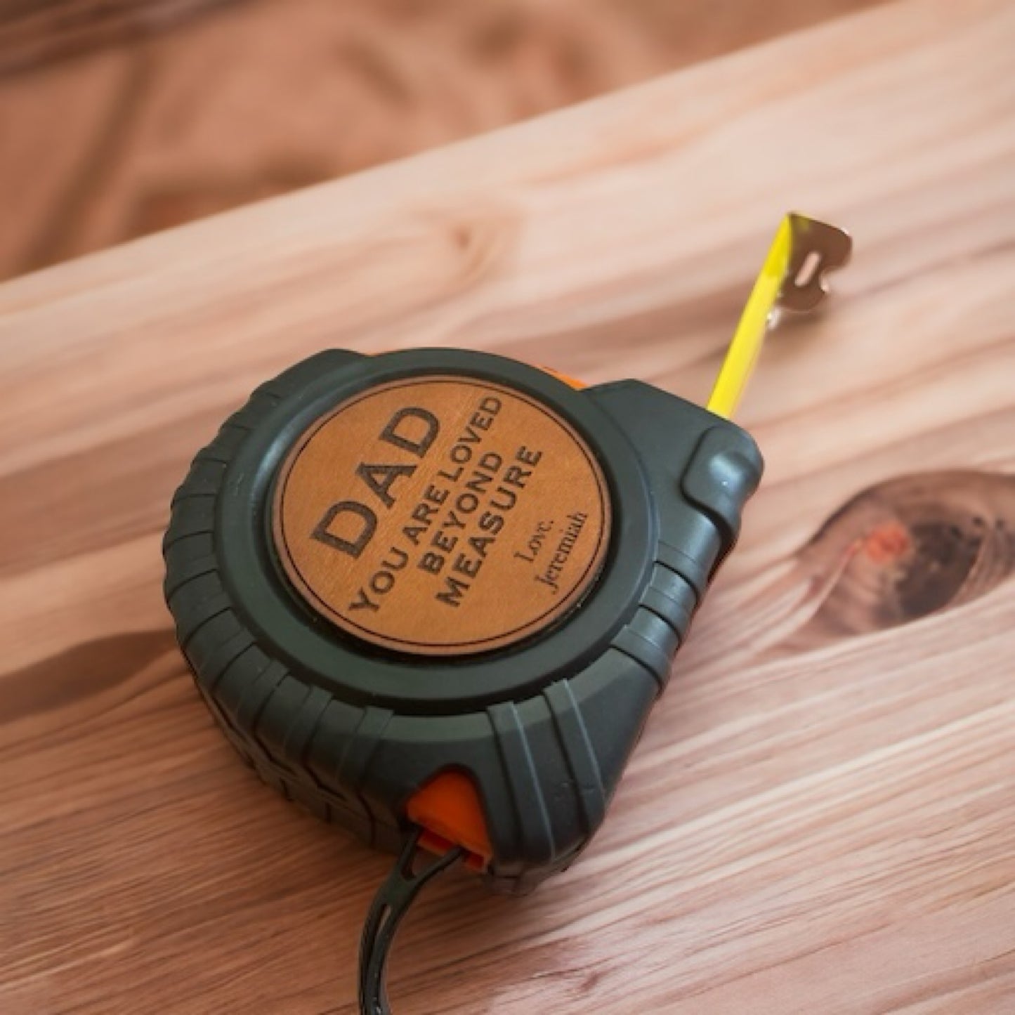 A personalized tape measure with a black and orange casing, featuring a leatherette patch with the inscription 'DAD - LOVED BEYOND MEASURE' and a yellow measuring tape retracting from the patch.