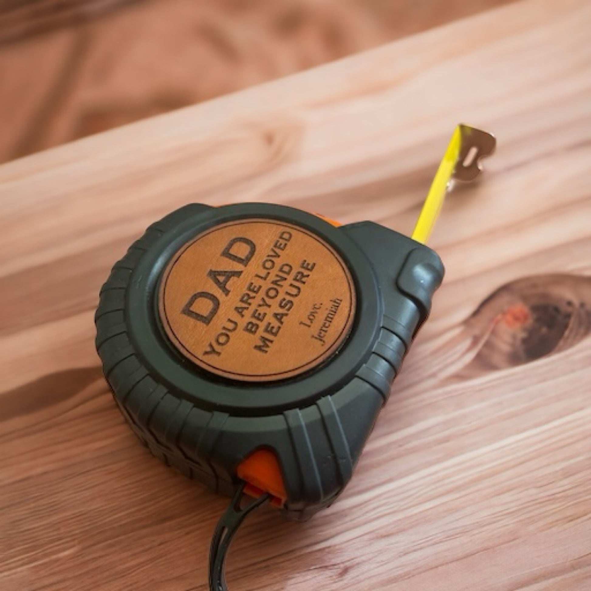 A personalized tape measure with a black and orange casing, featuring a leatherette patch with the inscription 'DAD - LOVED BEYOND MEASURE' and a yellow measuring tape retracting from the patch.