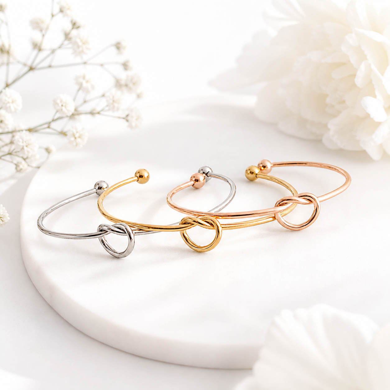 Three gold, silver, and rose gold bracelets on a white surface with flowers in the background.
