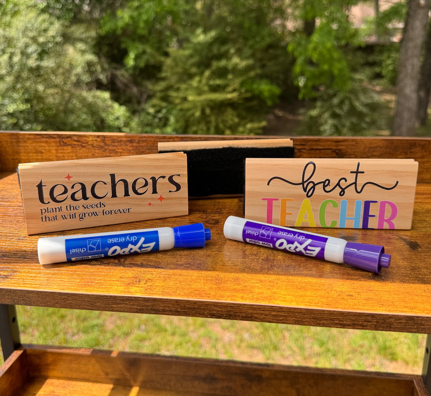 Wooden whiteboard erasers with 'teachers' and 'best teacher' text, along with Expo dry erase markers on a wooden surface with a natural background.