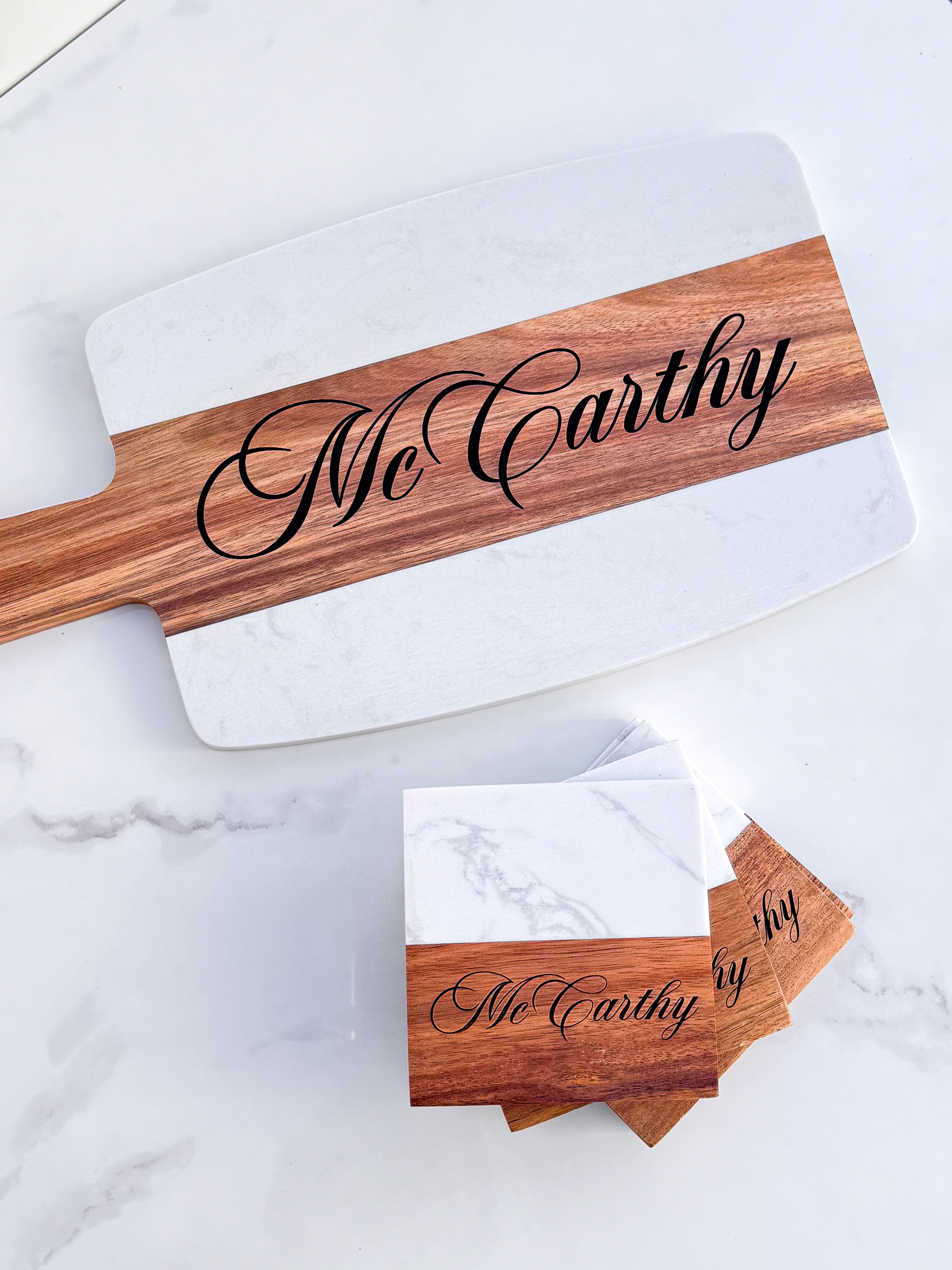 A personalized cheese board with 'McCarthy' written on it, alongside four coasters, all made of marble and acacia wood.
